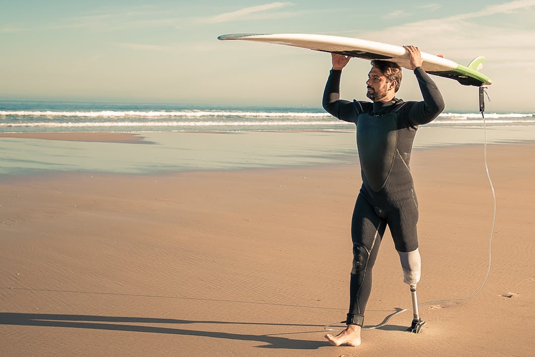 man in wetsuit with prosthetic leg walking on beach holding a surfboard