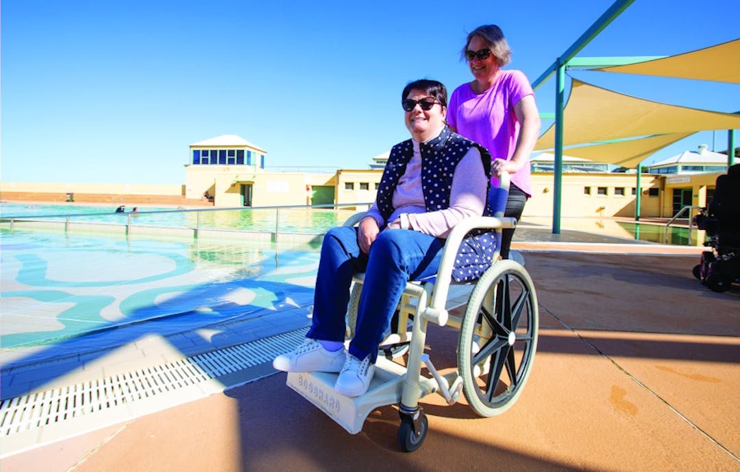 A woman pushes another woman in a beach wheelchair at Port Kembla Pool on a sunny day. Both are smiling.