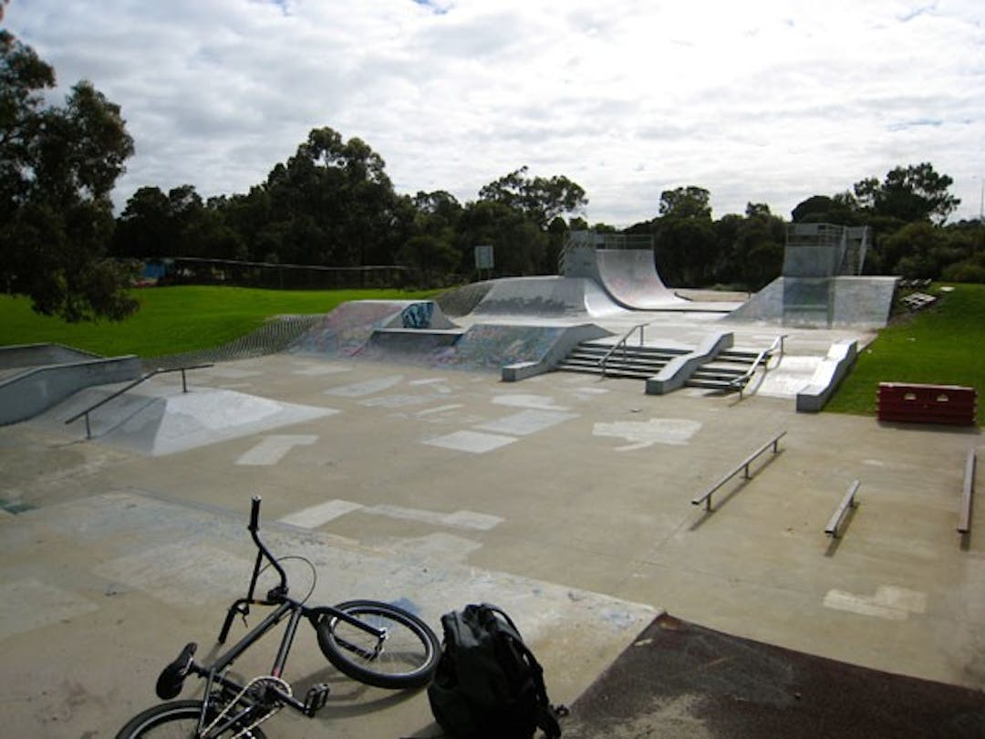 A concrete skate park with ramps and grind rails. A bmx bike and backpack are lying on the ground at the front.