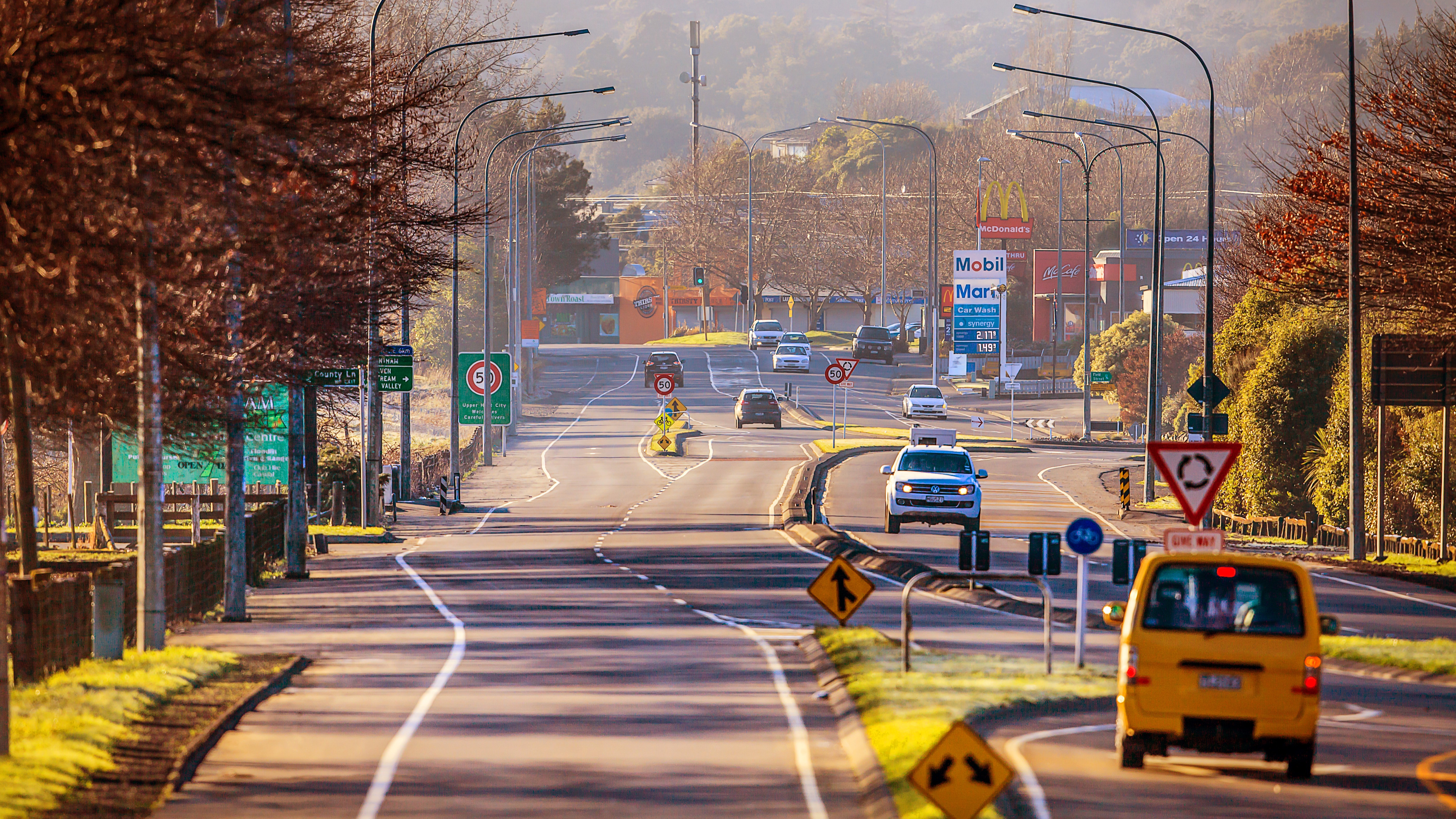 Long stretch of road with trees lining the left side of the road and vehicles on the right side of the road