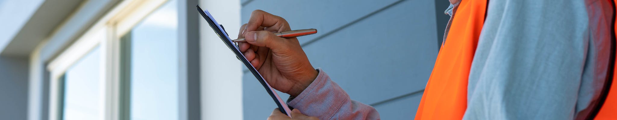 Person writing on a clipboard near a house, wearing a highly visible vest.