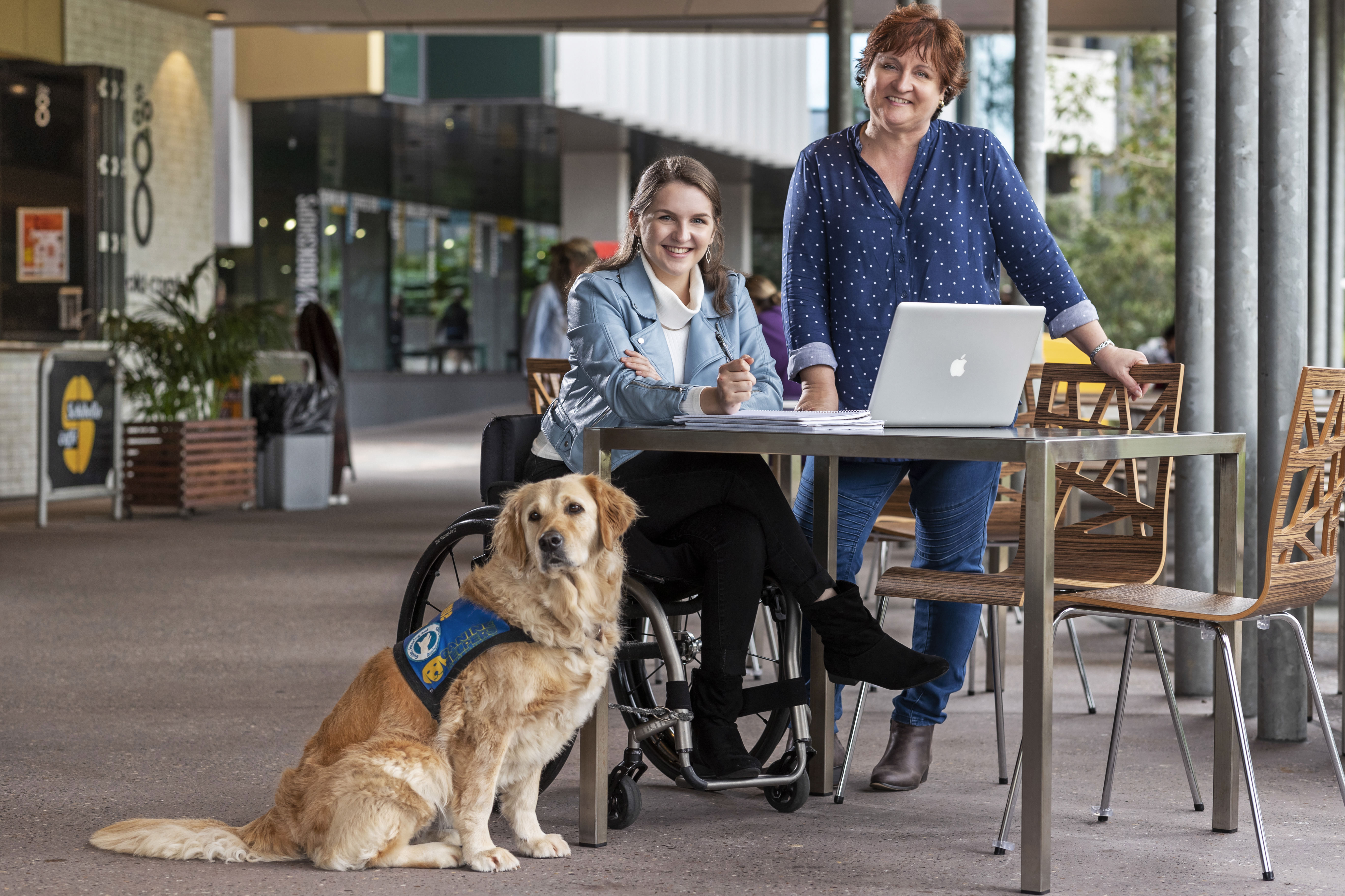 Student in wheelchair with service dog studying at table with staff member at university campus
