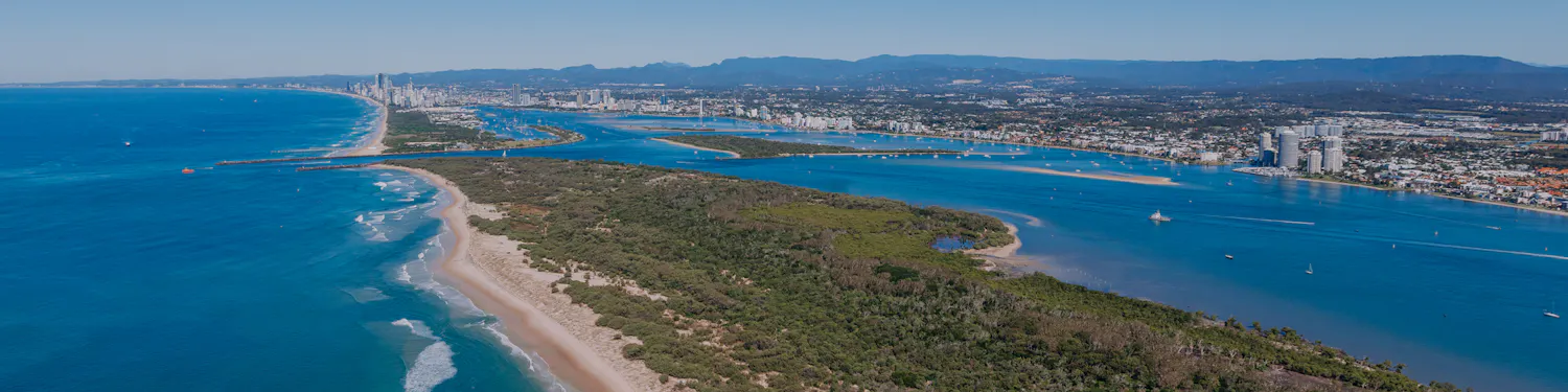 Aerial view of South Stradbroke Island showing the ocean beach, vegetated dunes, waterways, and the mainland skyline.
