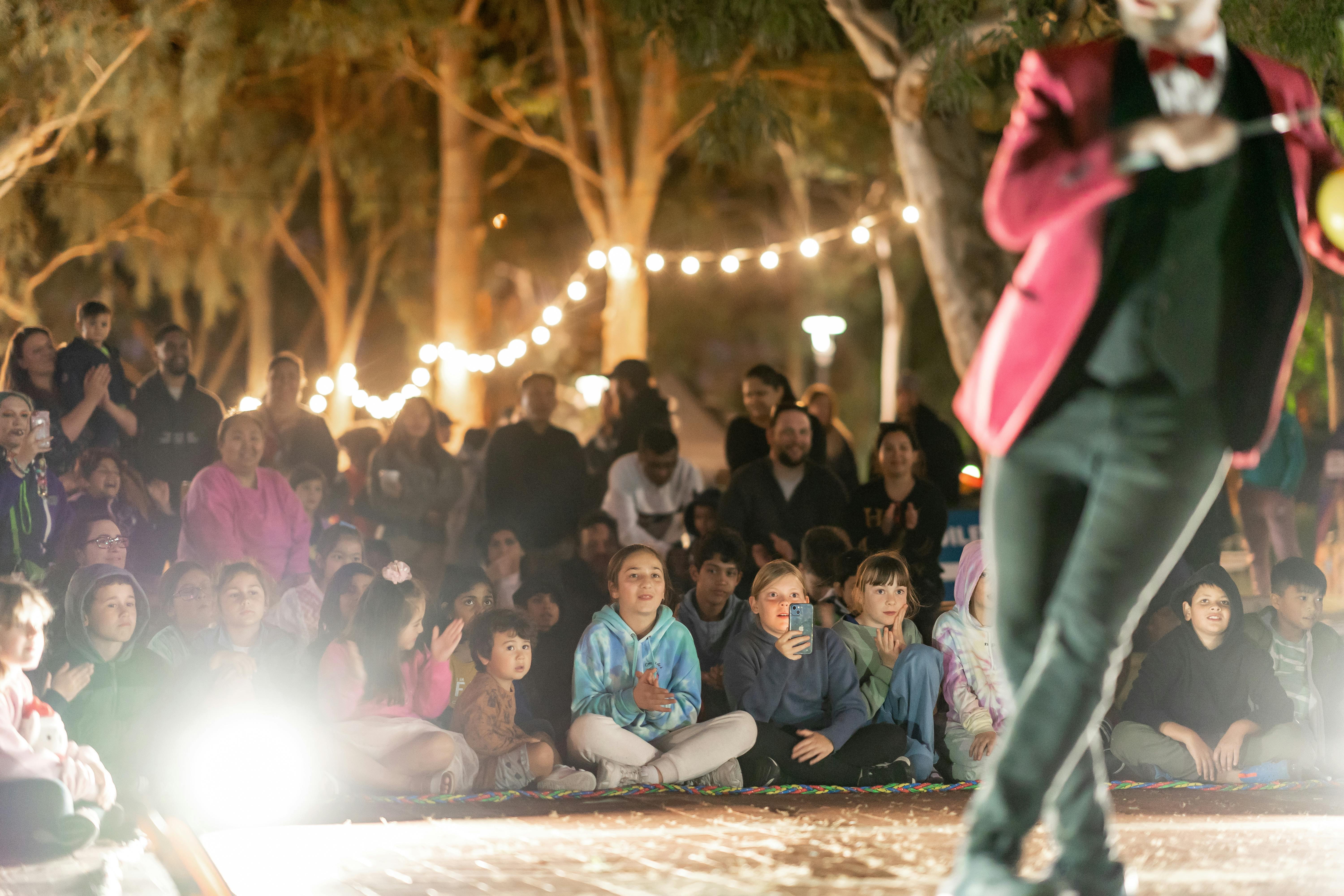 A performer in a red circus ring masters jacket dances to a large crowd sitting in front of trees and festoon lights.