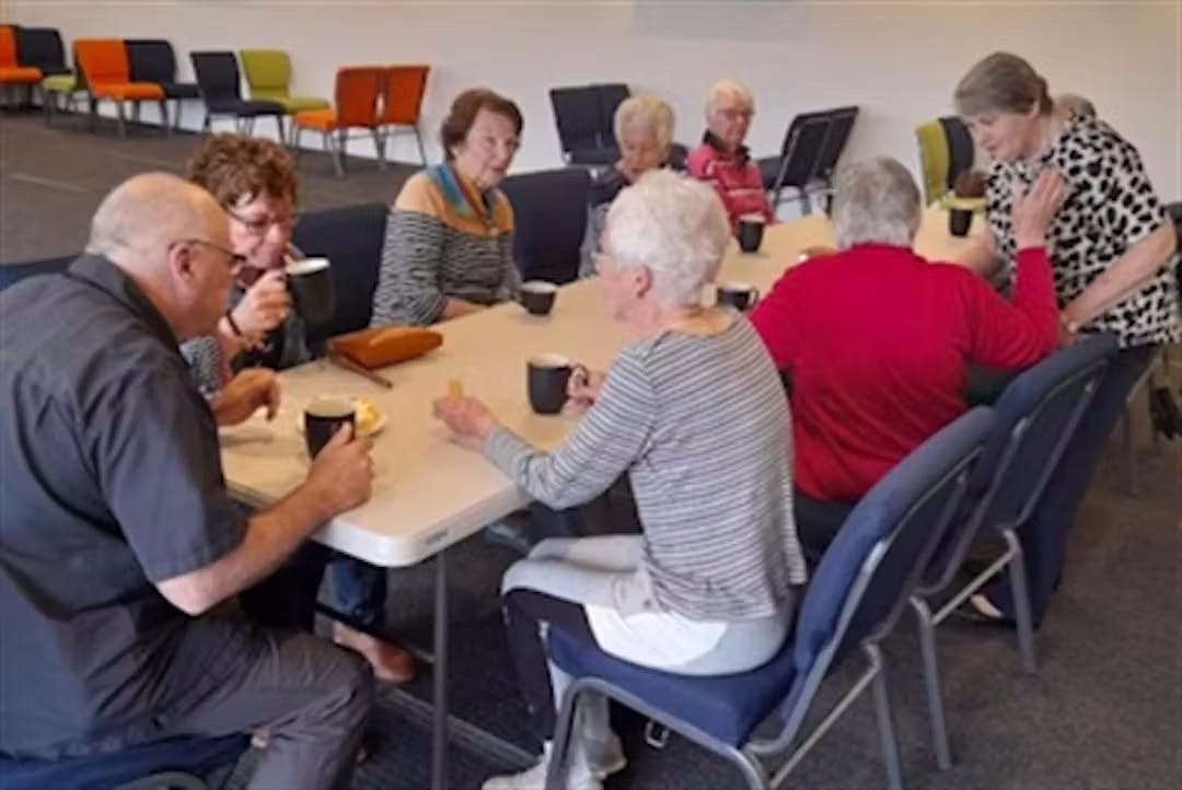 Older people sit around a table drinking hot drinks and talking