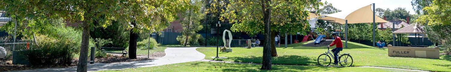 Tree and open space at Henry Codd Reserve with a playground in the background, a cyclists rides past on a pathway