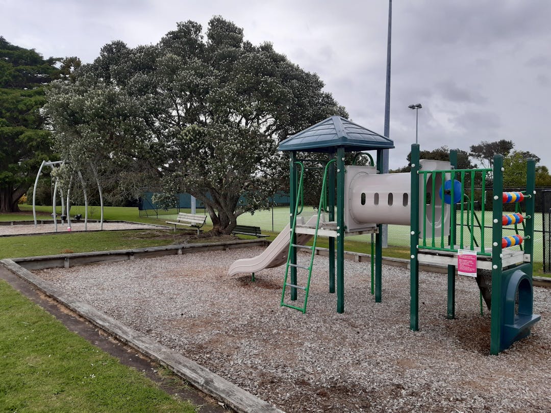 An image of a playground with a tower with a slide tunnel stairs and climbing frame, set in a wooden pit filled with wood chips. In the background is the grass of the park and the parking lot at the edge.