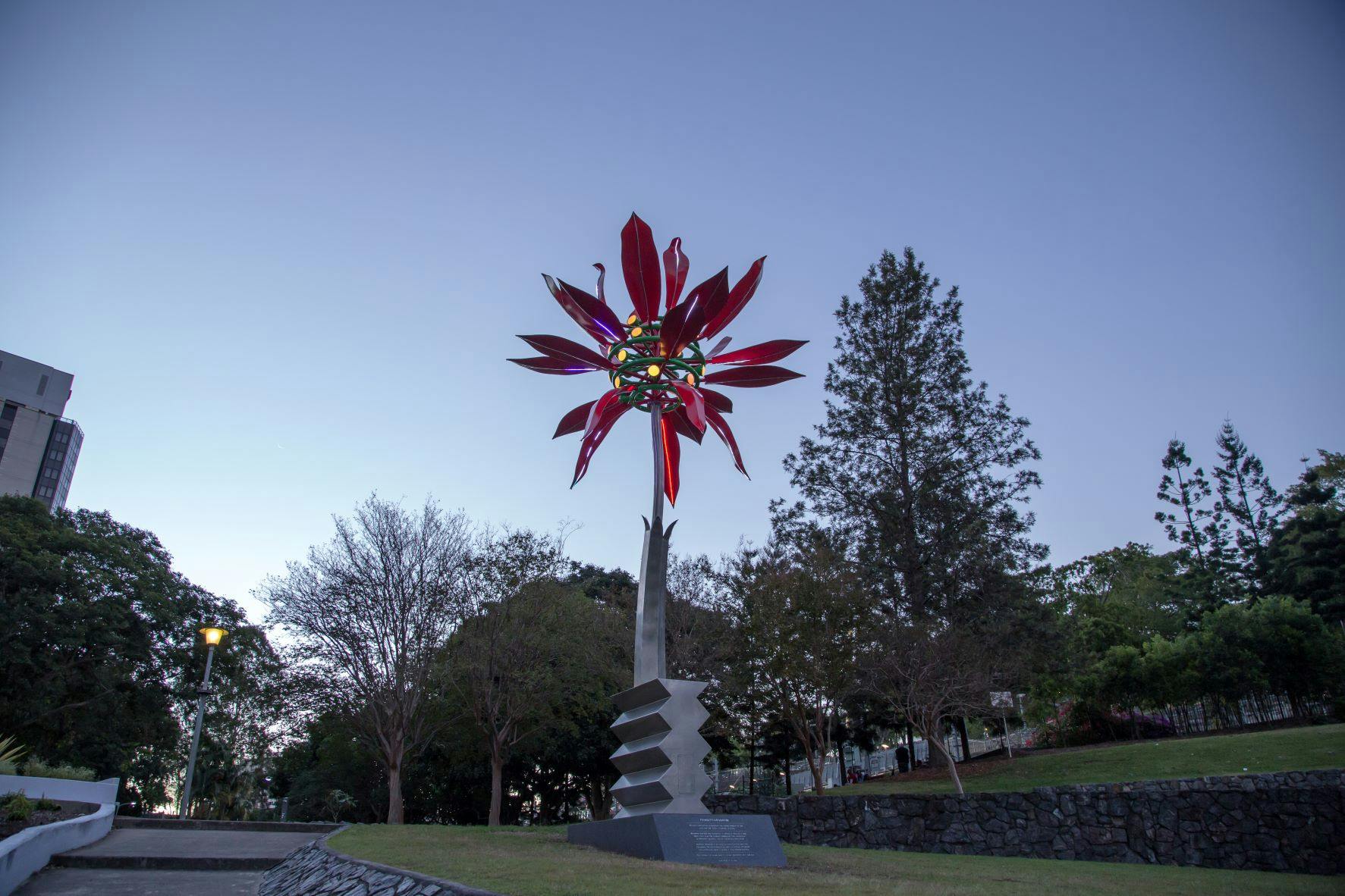 Image of Poinsetta Riverfire sculpture located in Emma Millar Place, Roma Street, Brisbane with trees and lawn in the background. 