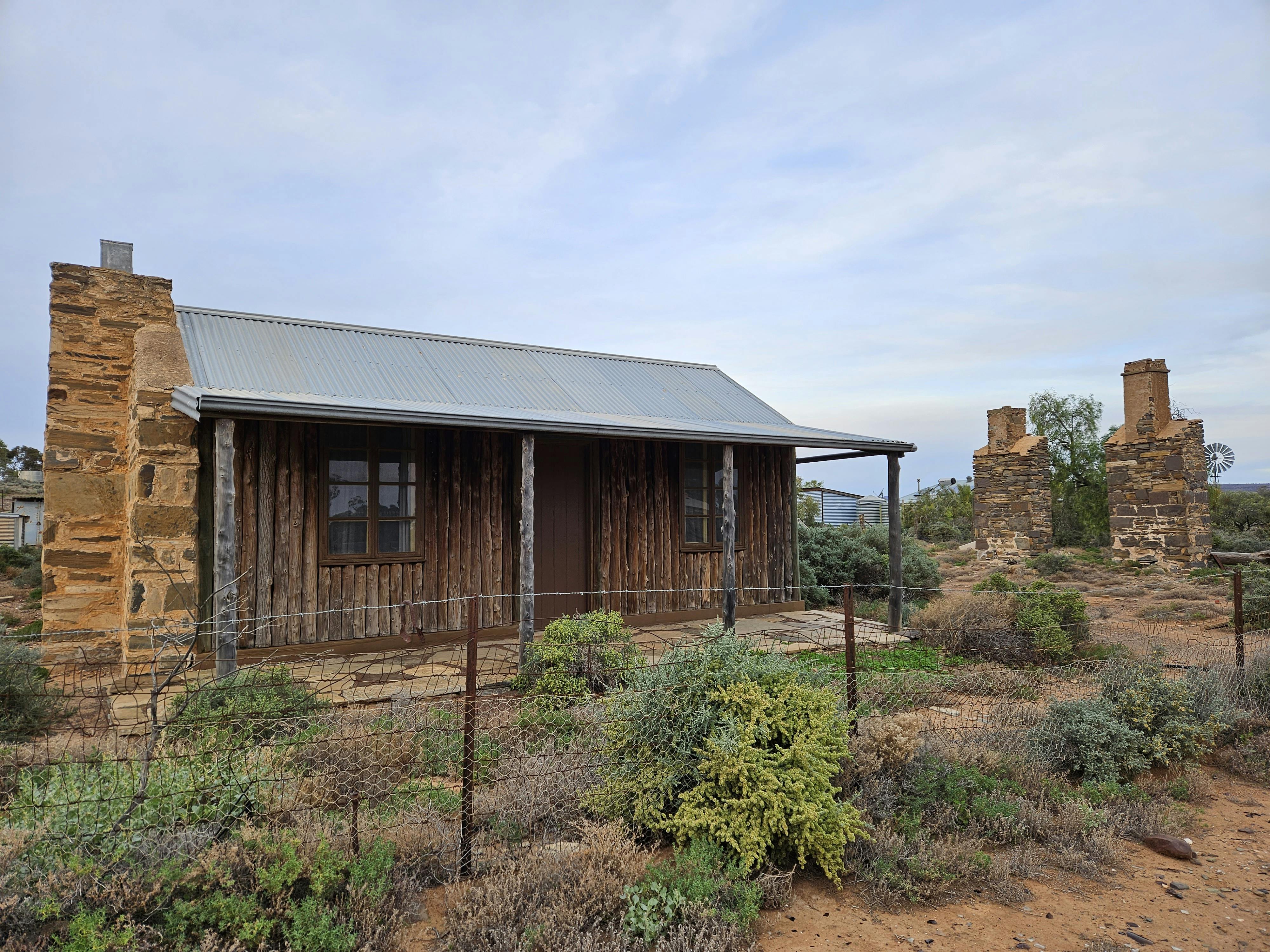 Historic cottage in good preserved condition, with stone chimney and rough-hewn timber front wall and verandah posts.