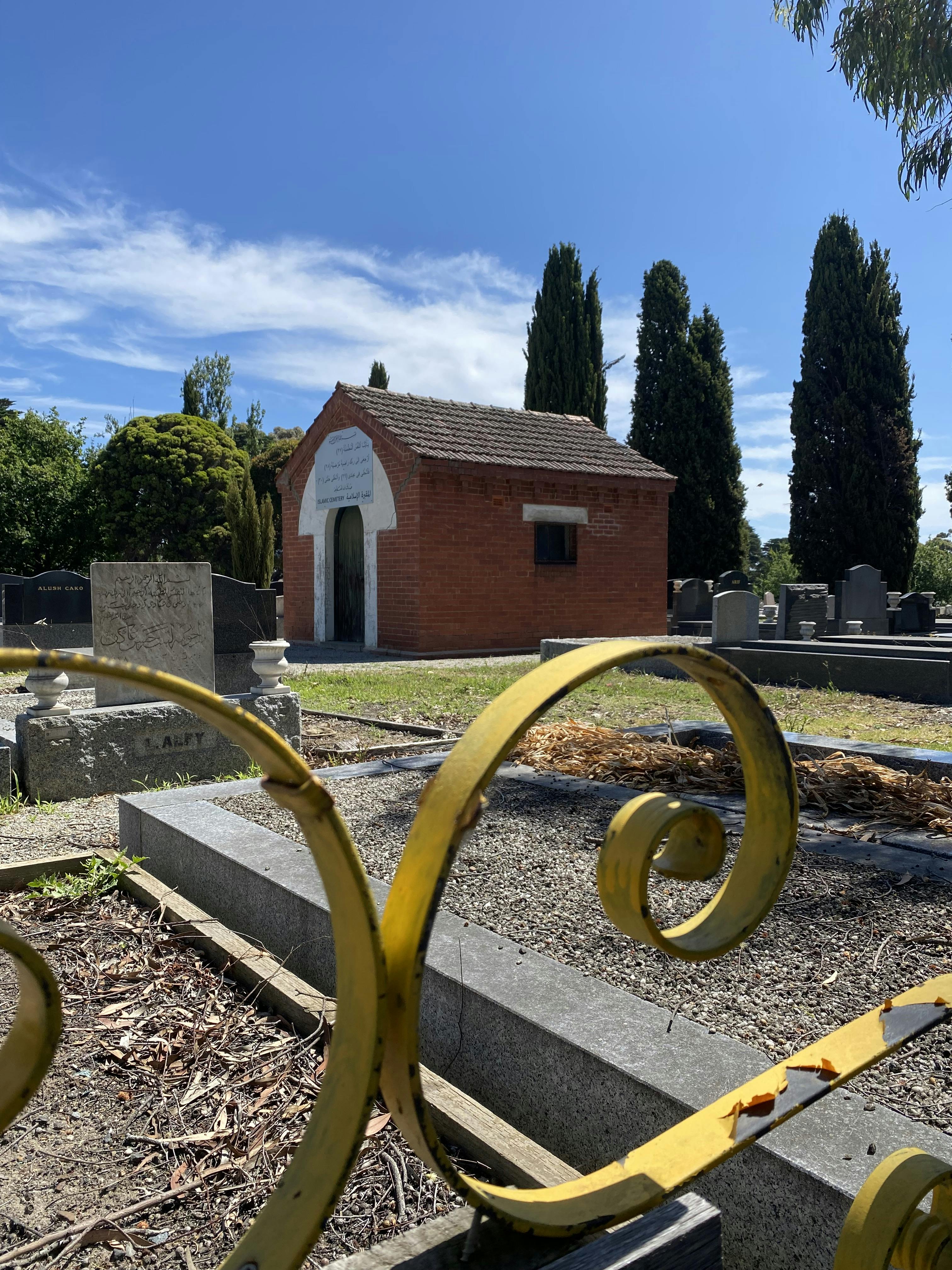 A red brick Islamic chapel sits with graves in the foreground and pine trees in the background. Curved yellow wrought iron is in the fore ground. 