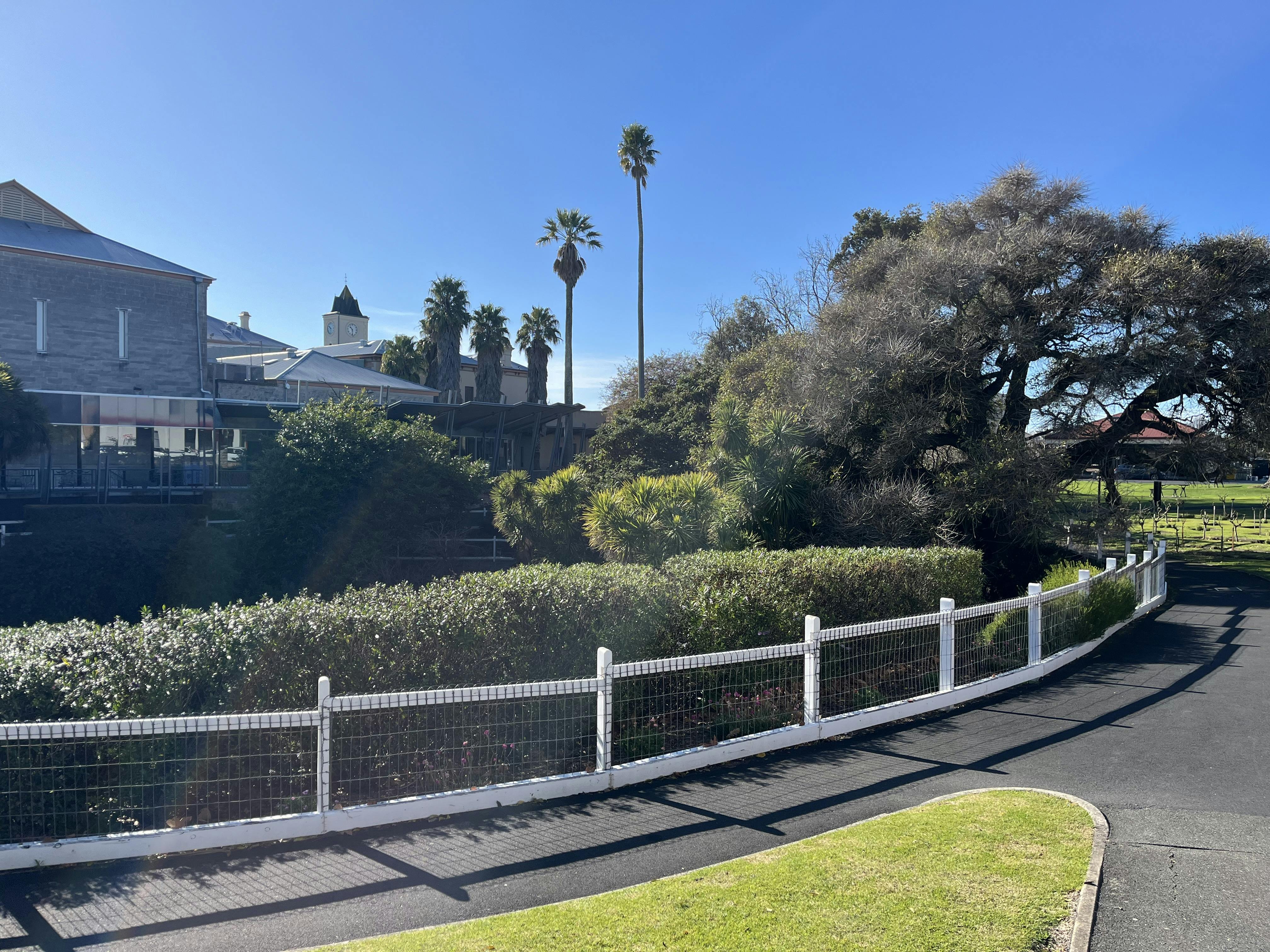 A low white fence surrounds a mature garden with stone buildings in the background