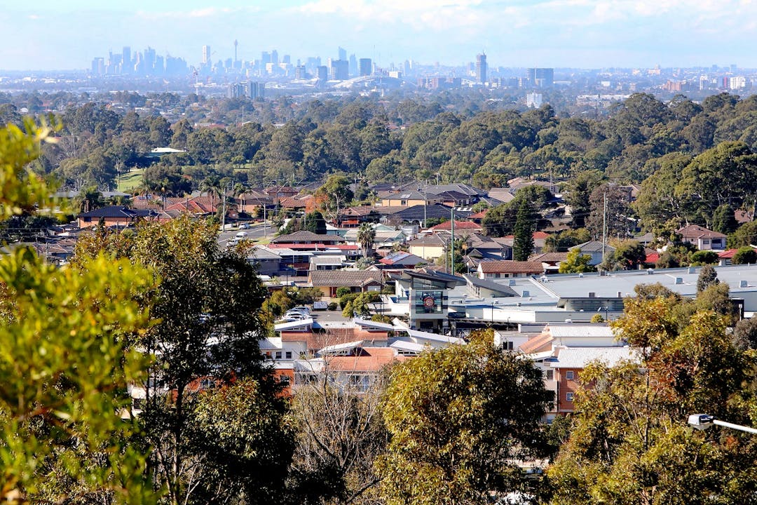 View of housing in Pemulwuy from Prospect Hill, with trees amongst the houses and skyscrapers in Sydney CBD in the background.