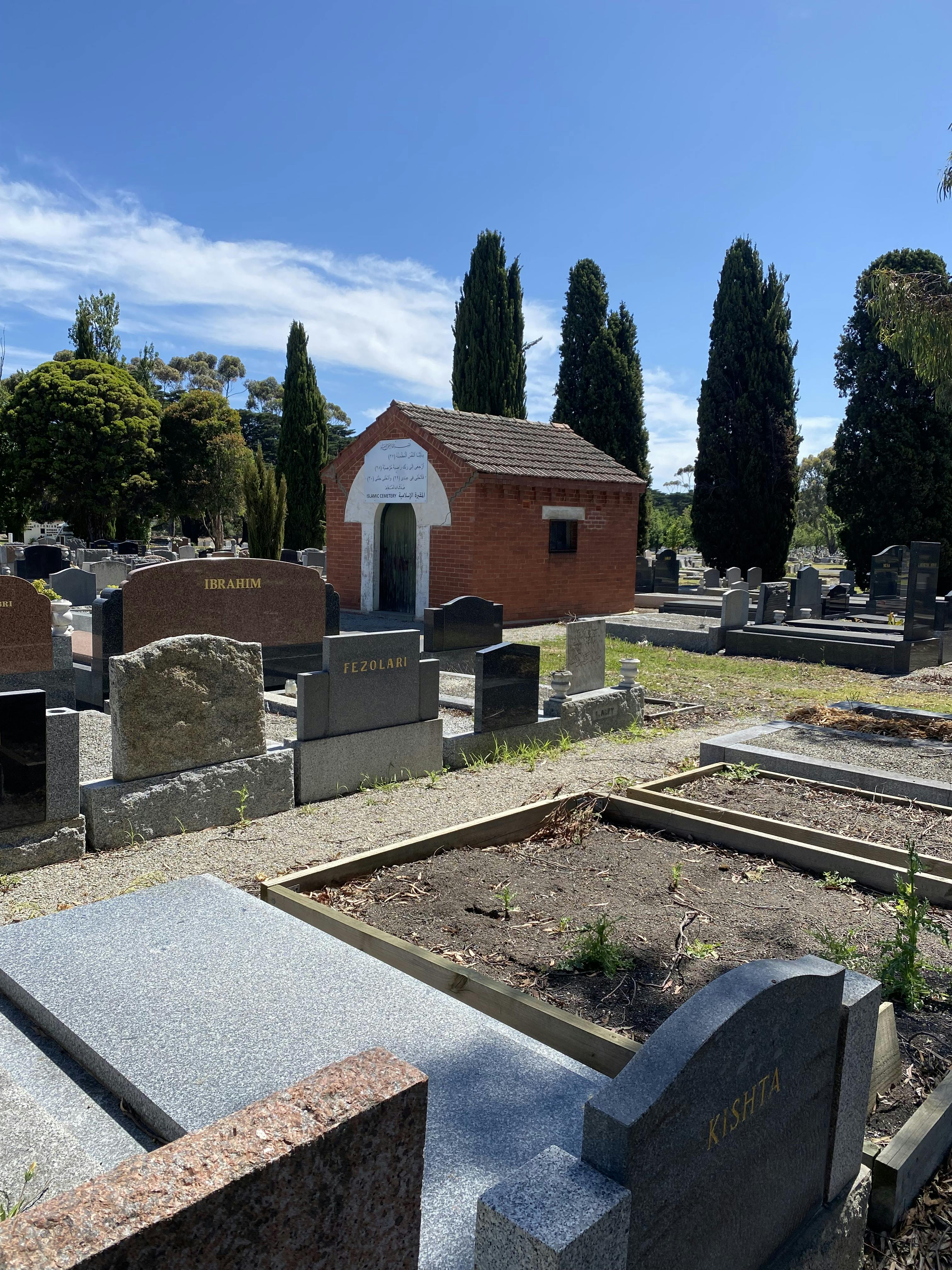 A red brick Islamic chapel sits with graves in the foreground and pine trees in the background