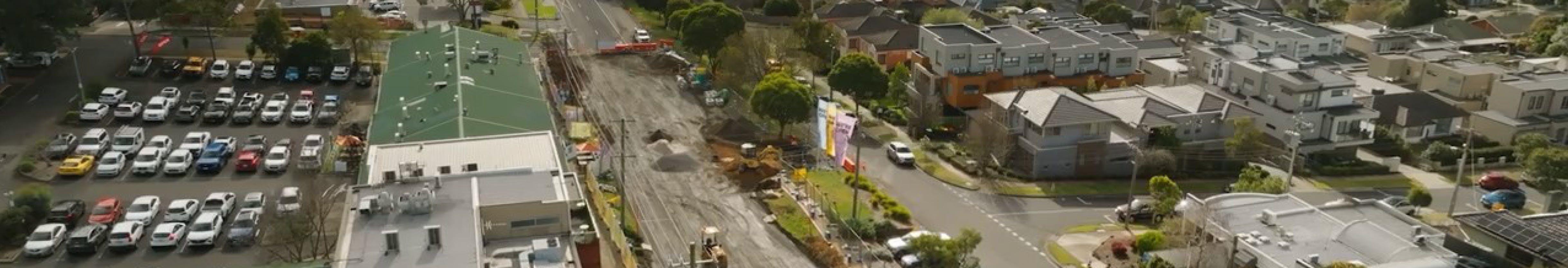 Aerial view of suburban area with a construction site, green-roofed building, parked cars, homes, trees, and roads.