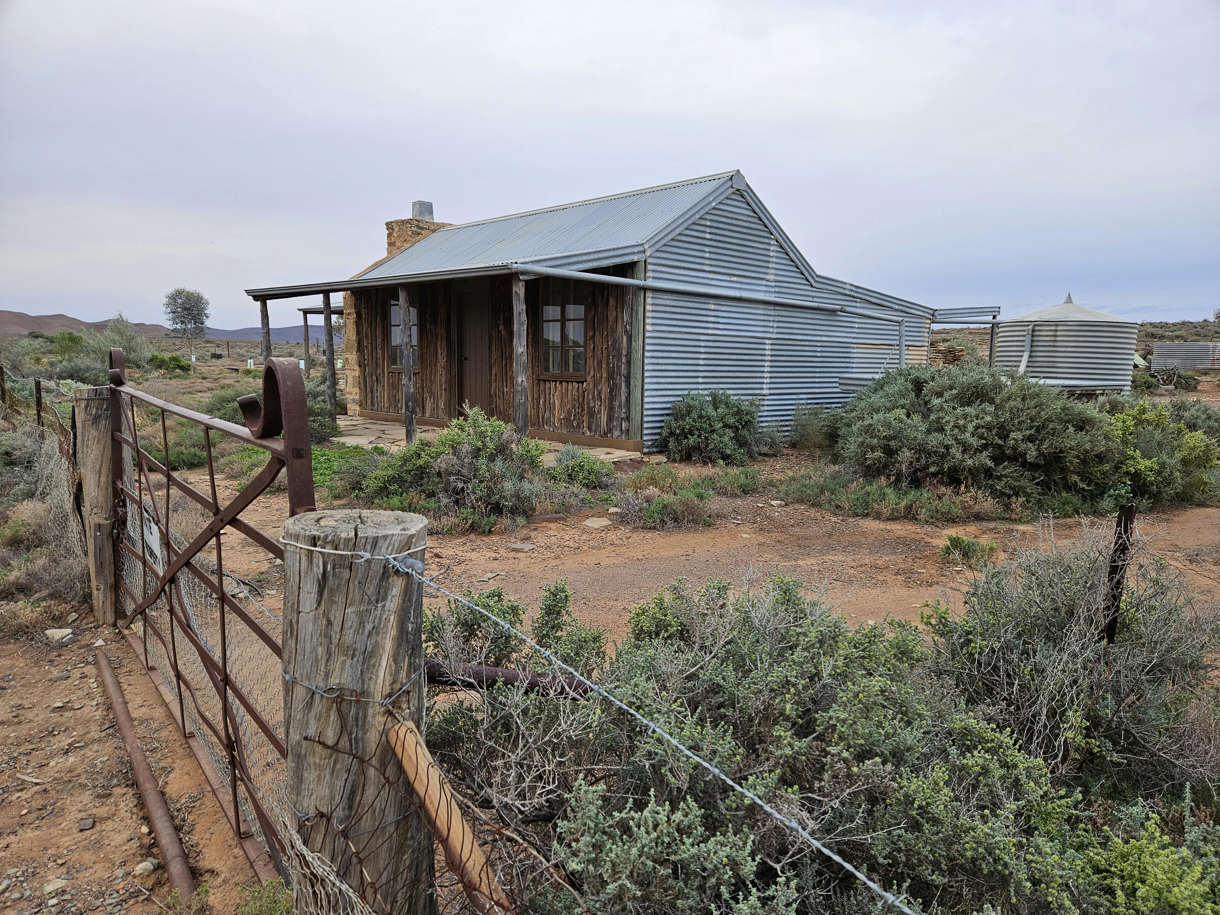 Rustic cottage with stone chimney, walls of timber slats and corrugated iron, with corrugated rainwater tank.