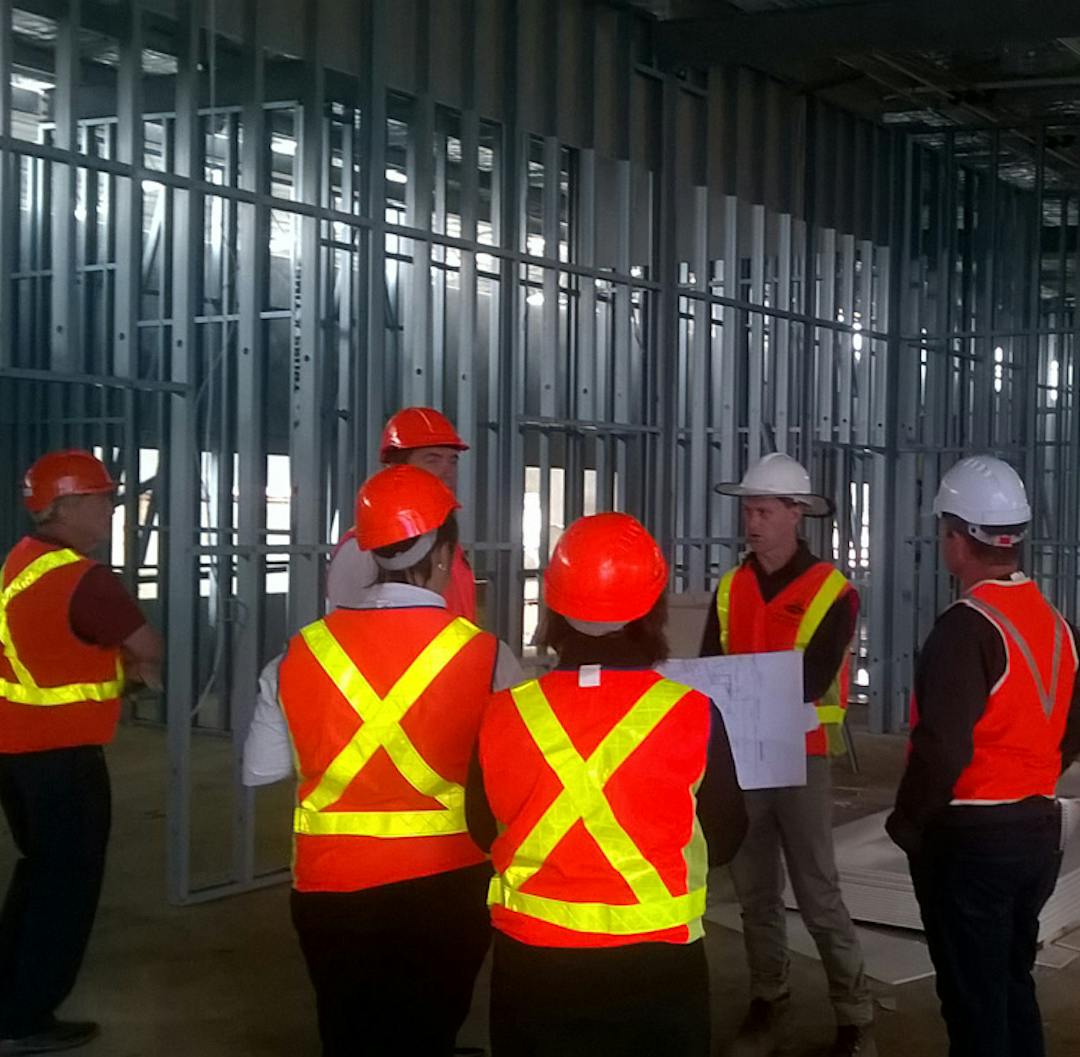 Six workers wearing high visibility vests and hard hats are standing inside a stadium being constructed.