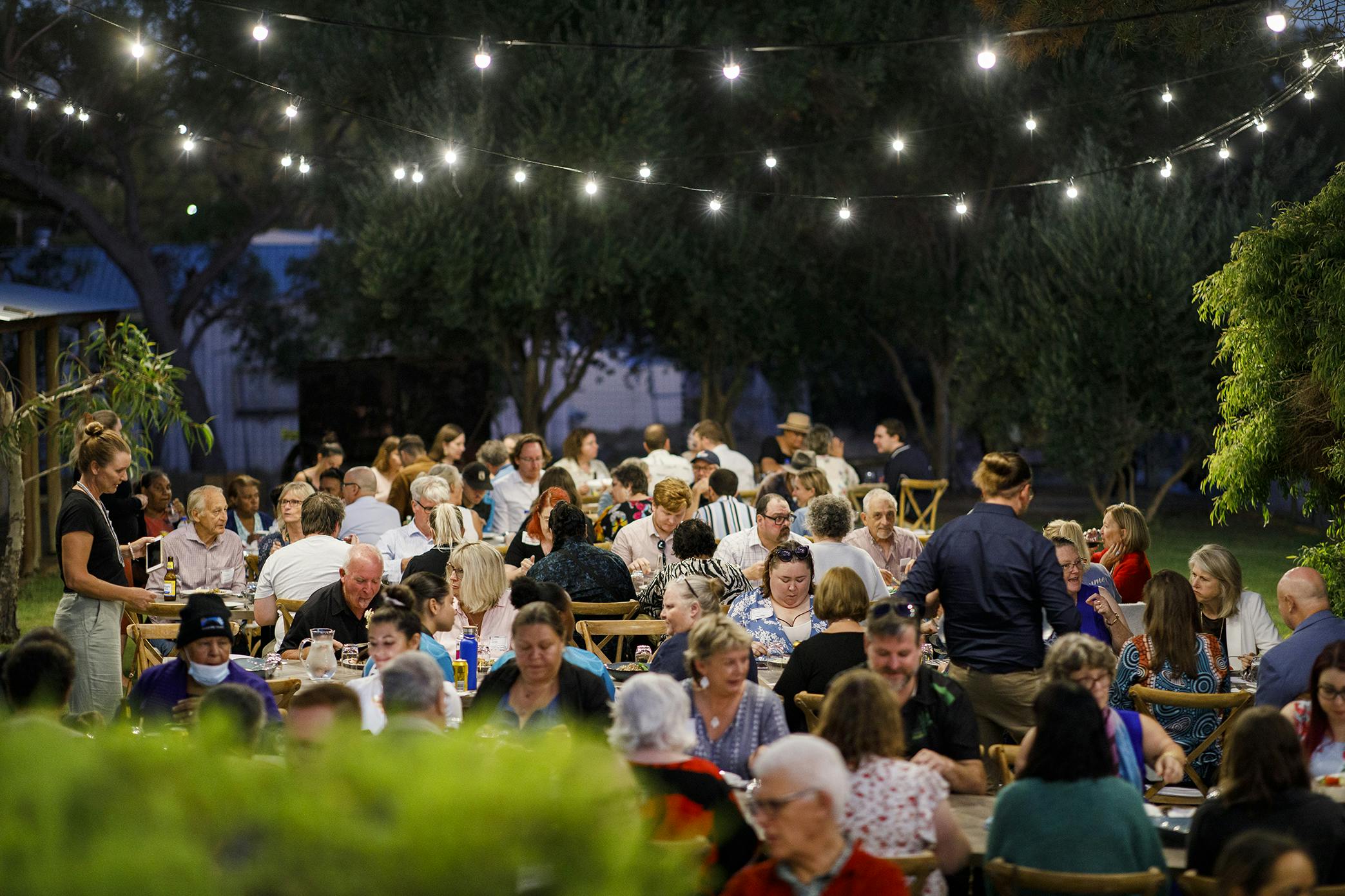 A large group of community members sit at long tables, in a garden, under festoon lighting. 