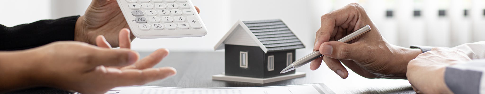 A photo of two people's hands opposite one another at a desk. One person is holding a calculator and one is holding a pen.