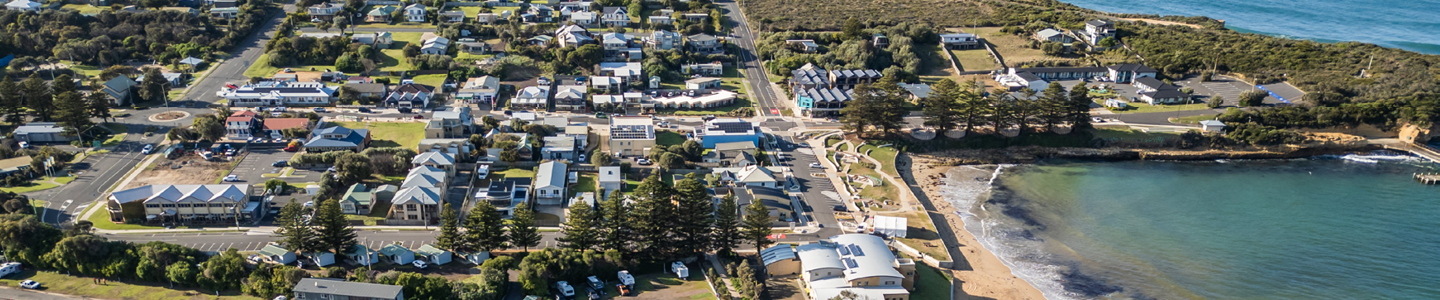 Aerial view of Port Campbell township and foreshore, showing streets, homes, shops, Norfolk pines, beachfront parking and bay