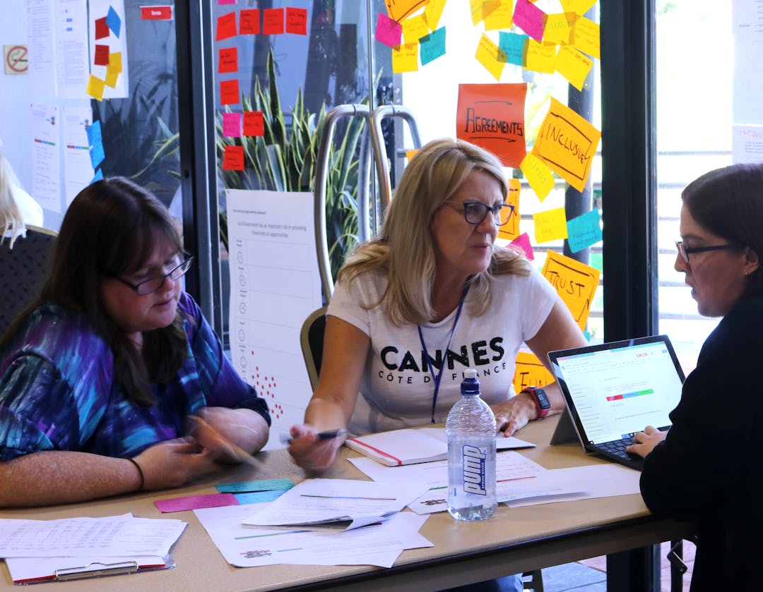 Three women sit at a table covered in papers and a laptop. They are in discussion. There are colourful post it notes on the window behind them.