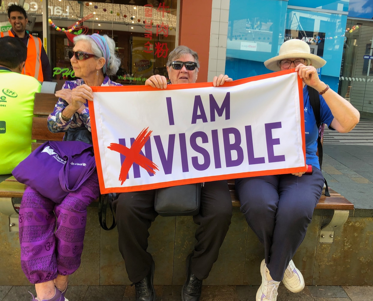 Three older women sitting on a bench holding a banner that says I am invisible with the letters i and n crossed out.