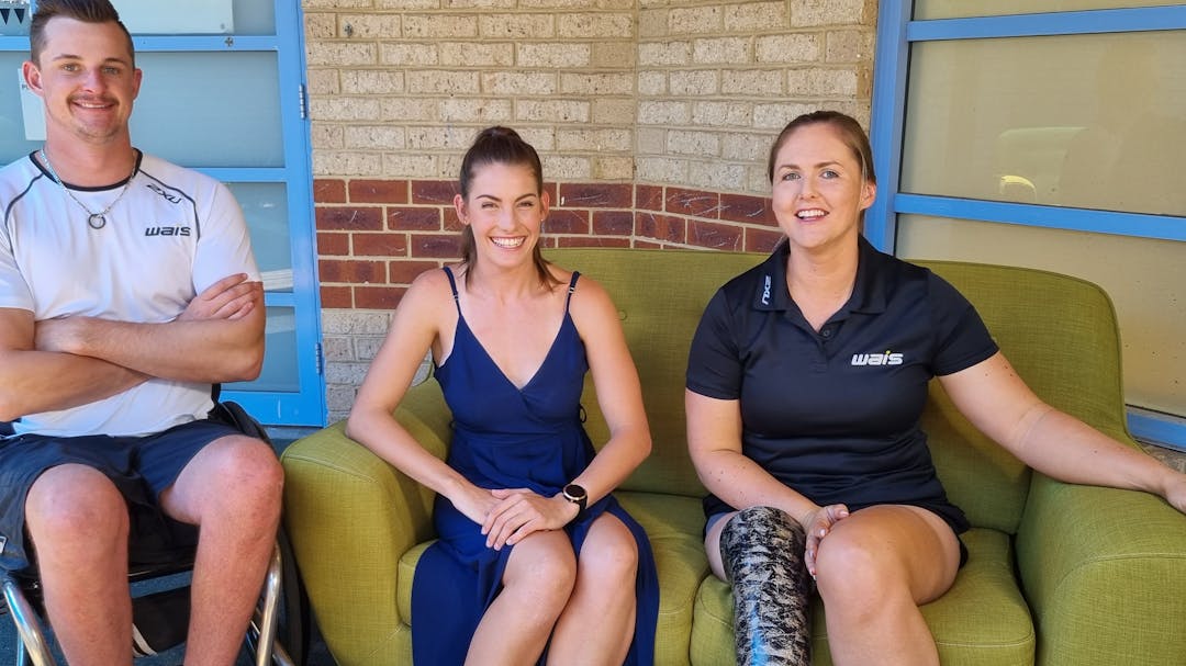 Three smiling young people with disability seated: a man in a wheelchair in sports kit and two women on a green sofa.