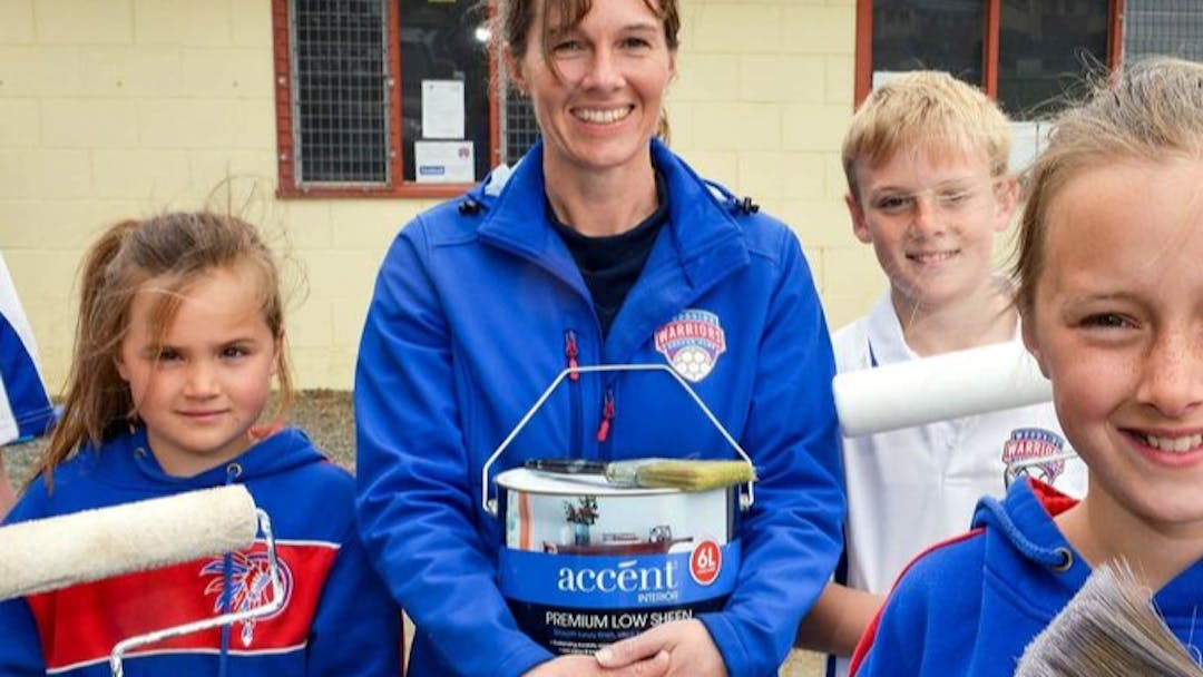 A woman and 5 children smiling in front of clubrooms holding paint and painbrushes