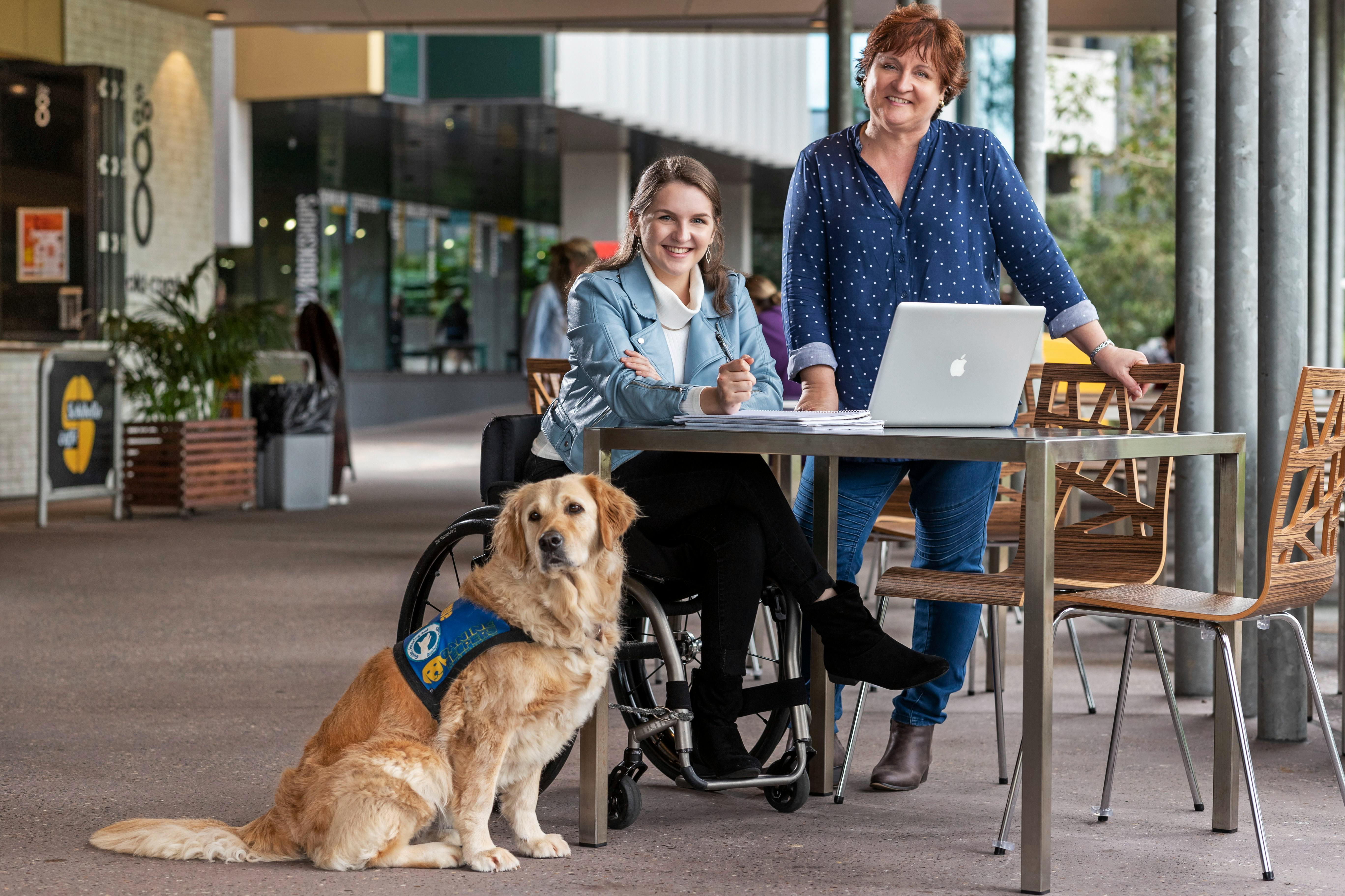 Student in wheelchair with service dog studying at table with staff member at university campus