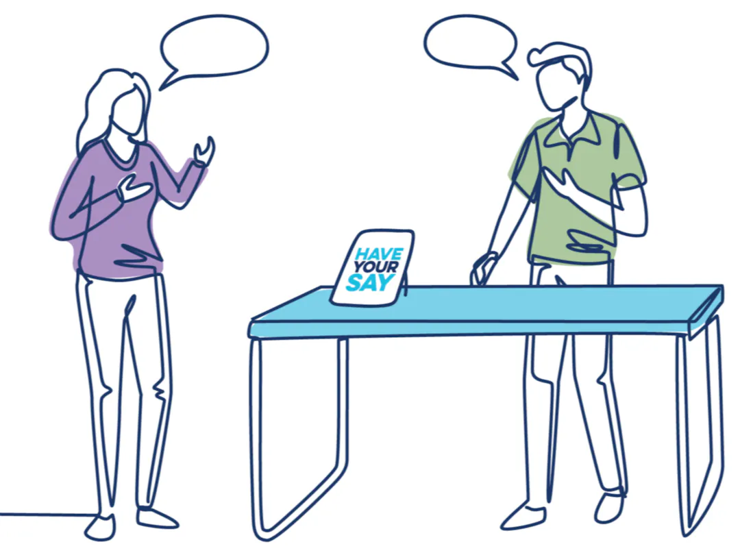Two people speaking at an in-person community consultation with a sign on the table that encourages sharing feedback.