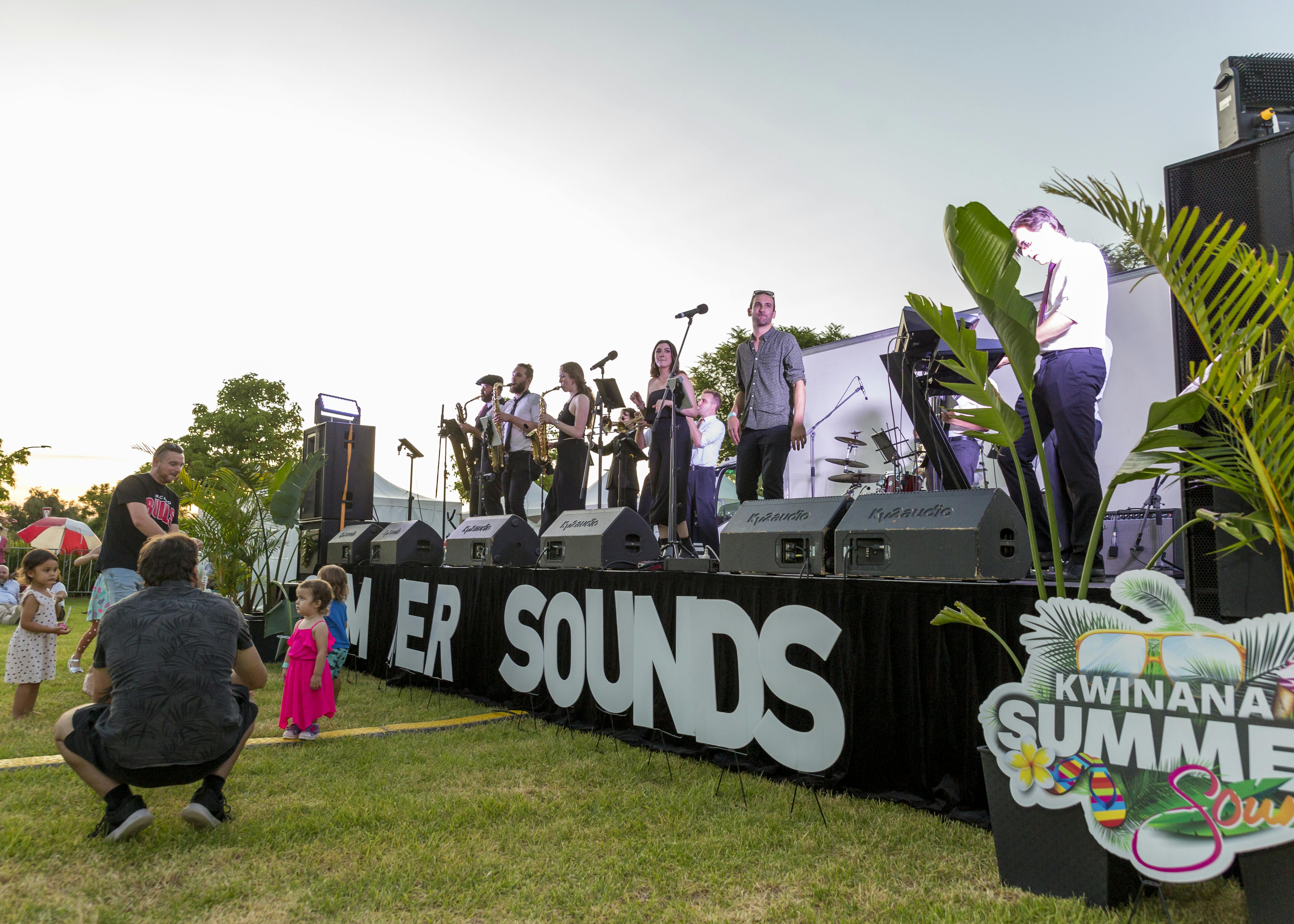 A 9 piece jazz band plays music on an outdoor stage with signage that says Summer Sounds  in the foreground.