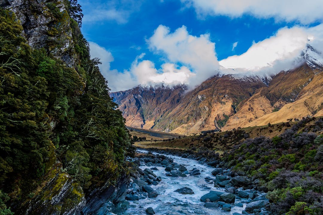 A scenic Otago landscape. There is a tree-covered mountain close to the viewer on the left, and a distant snow-covered mountain on the right. These two mountains are separated by a river which runs through the centre of the image.