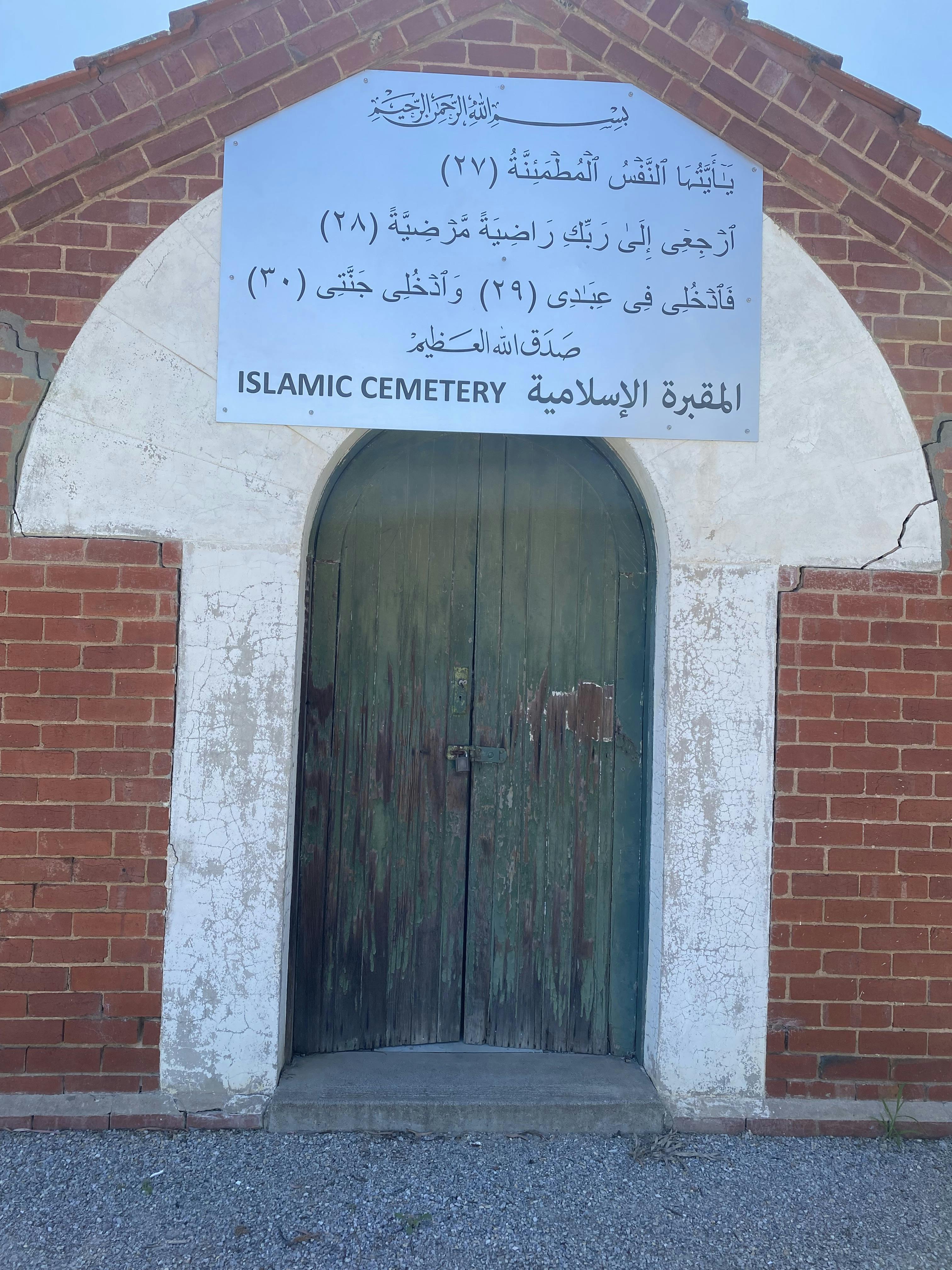 A faded sign with Arabic text is fixed on the red brick chapel with white rendered archway and green doors. 