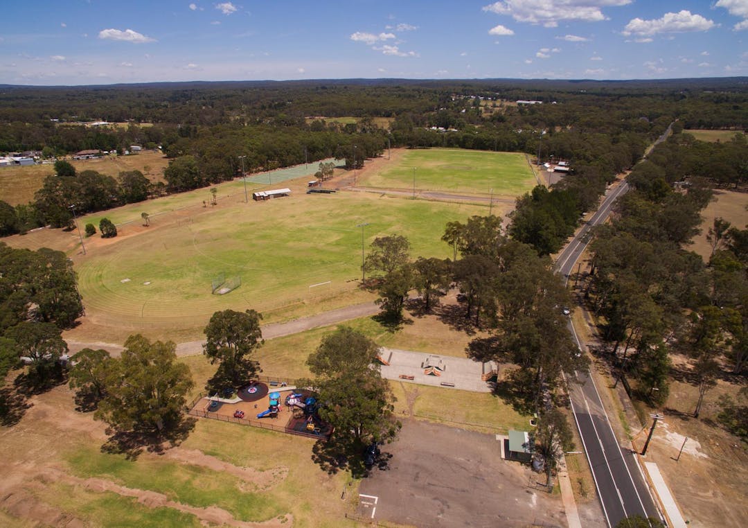 Aerial shot of Tahmoor Sportsground showing two sports fields, netball courts, play equipment, skate park, trees, and amenities.
