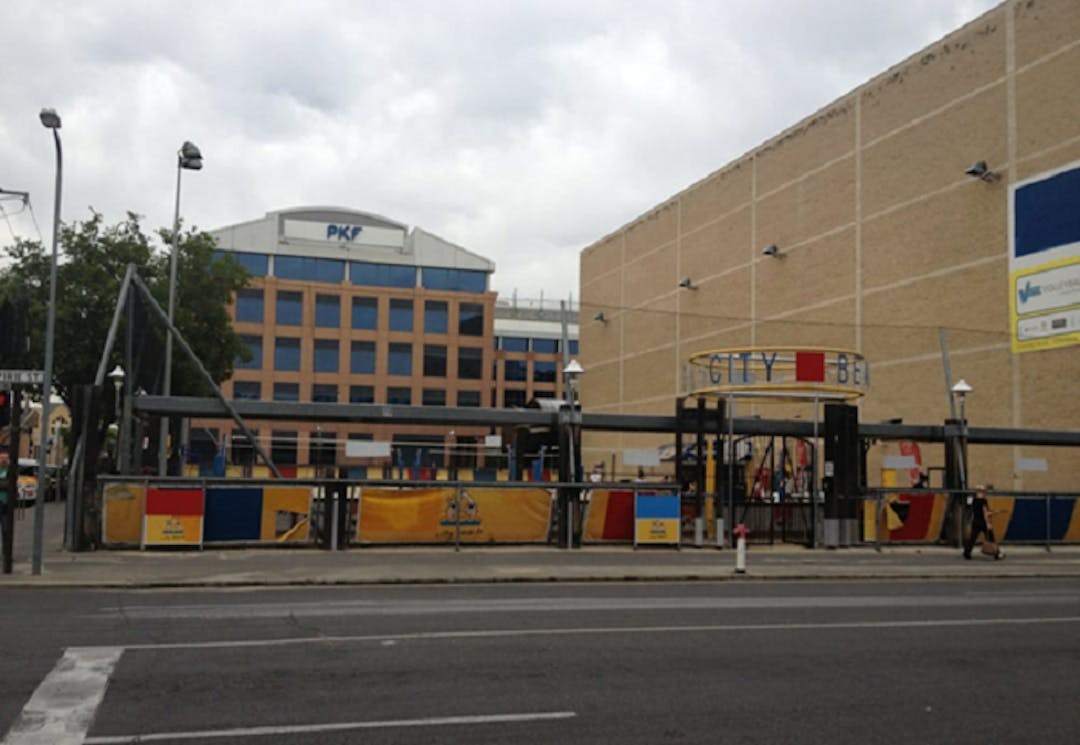 View of Pirie Street Volleyball Courts from Pirie Street. You can see the fencing around the volleyball courts, which extends to the pavement on the south side of Pirie Street and the entrance to the courts with 'City Beach' written above it is clearly visible. Also visible in the background is a building with large 'PKF' signage.