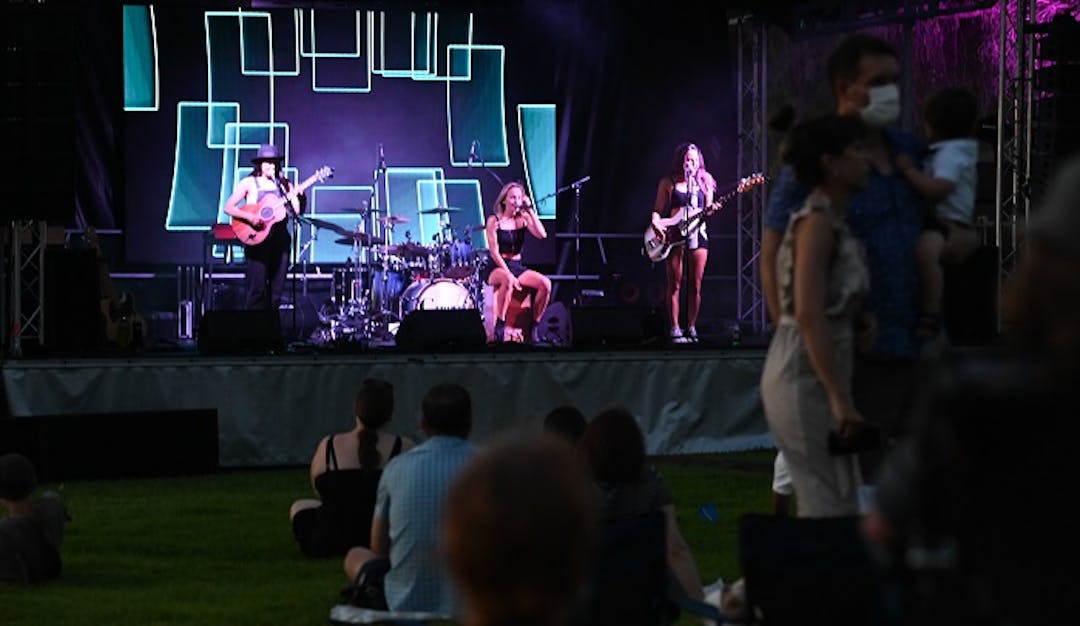 Band on stage at Rymill event; Purple/turquoise backdrop. Guitarist left; drummer back centre; singer sat front; bass right.