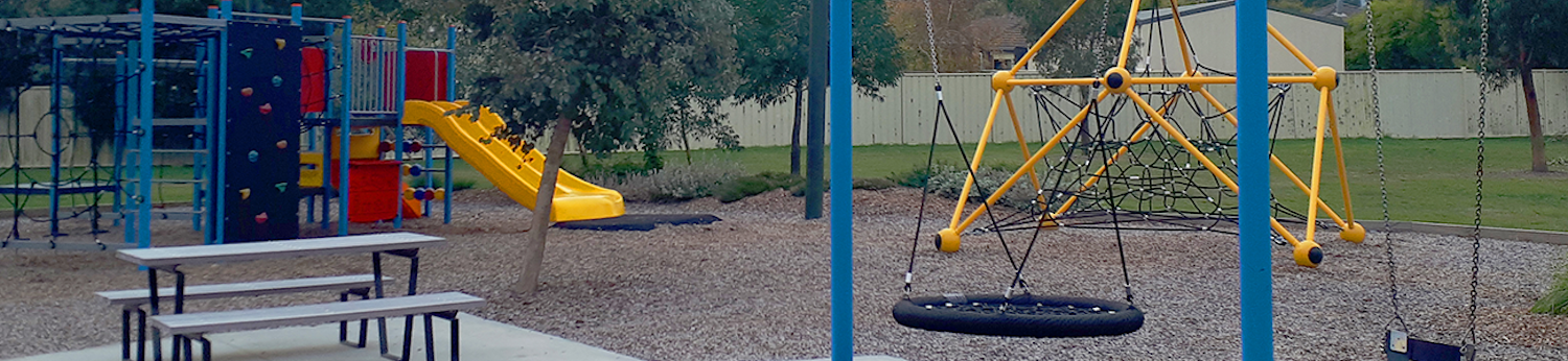 Playground with shade sails, swings, a table, climbing equipment and a slide with fence and houses in the background