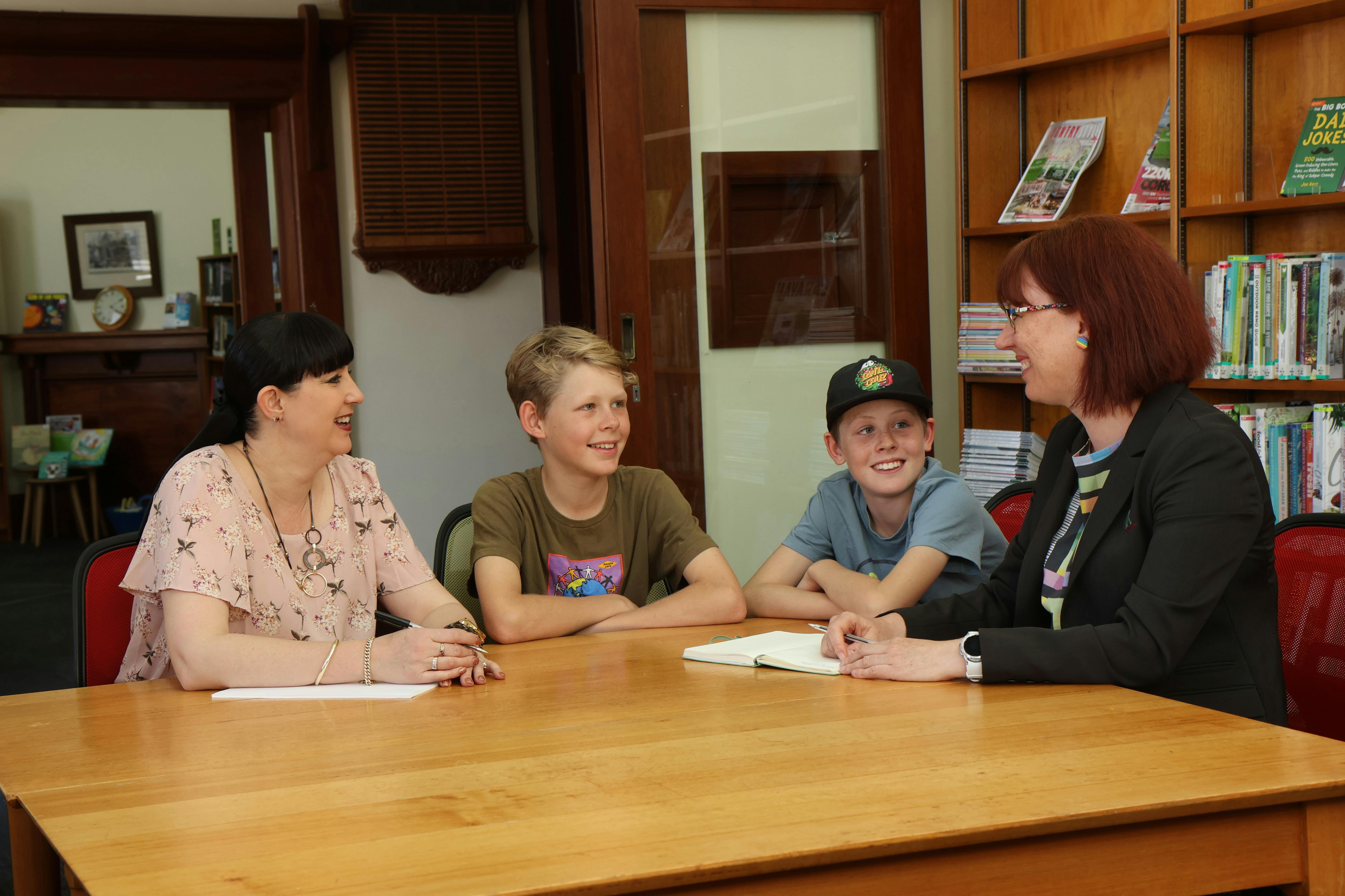Two Barossa Council staff members having a discussion to two school aged boys around a table in a library space