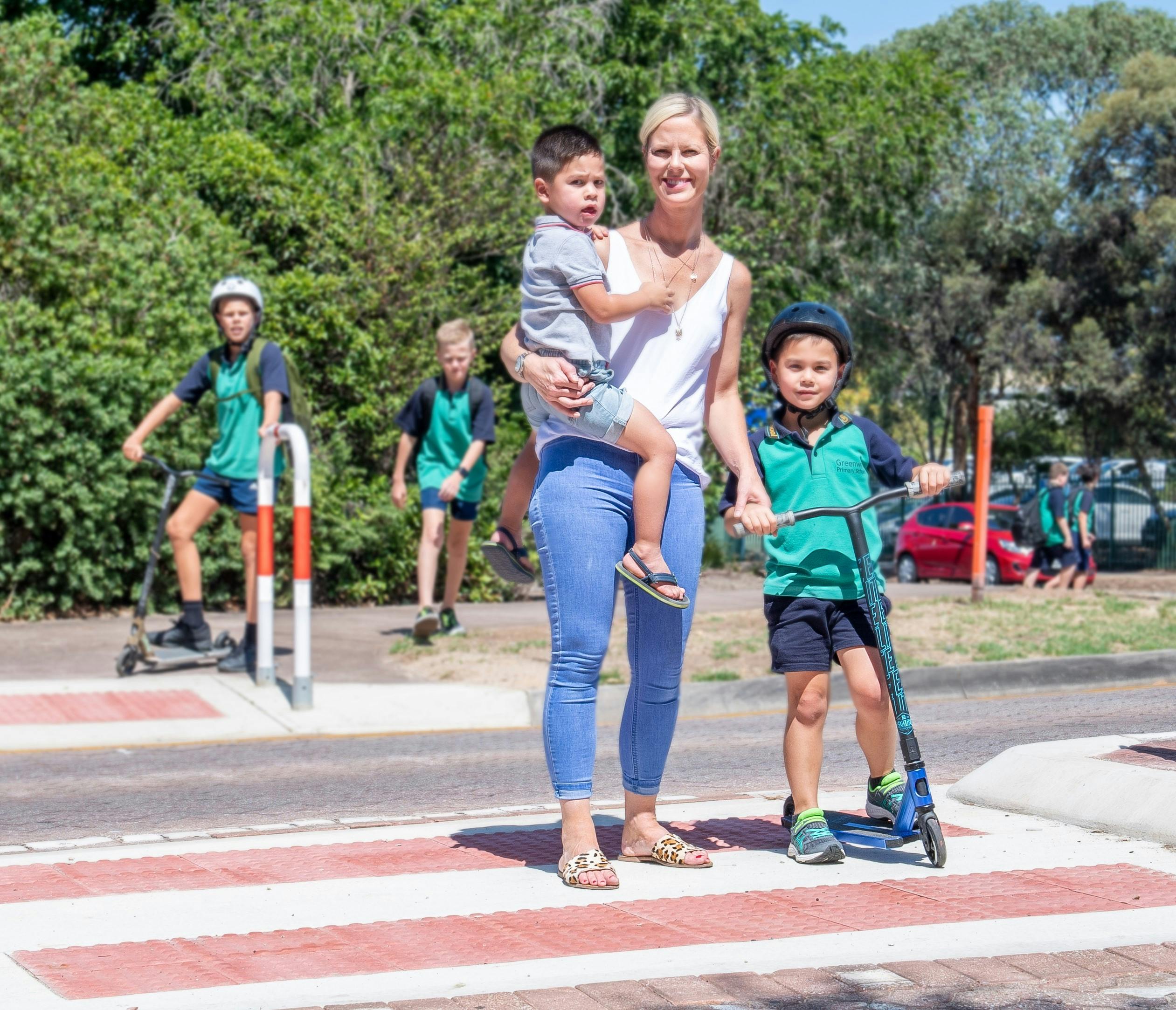 Mum holding children and another child on scooter using pedestrian crossing