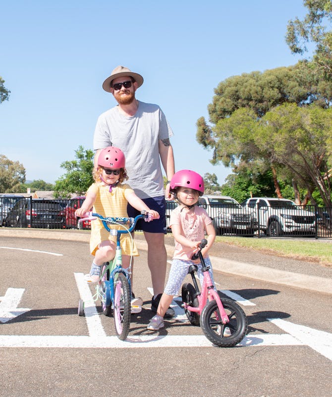 Man standing behind two young girls on bikes. The girls are wearing pink helmets and smiling at the camera