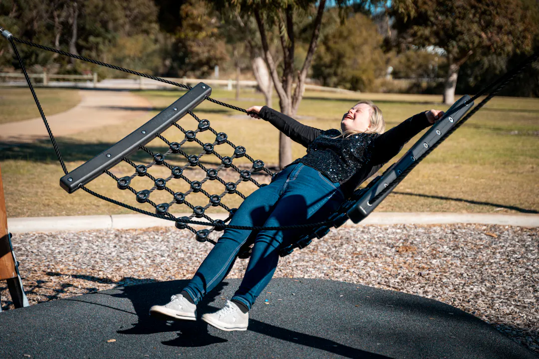 Young adult smiling while relaxing on an inclusive net swing at an outdoor playground in a park.