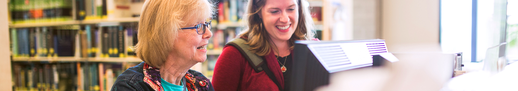 2 ladies sitting in front of a computer in a library with books behind them