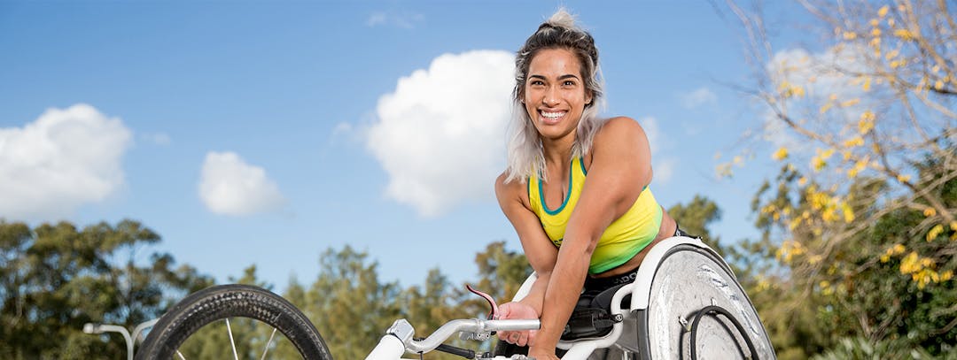 This is a decorative image. Madison De-Rosario, a young woman outside in a racing wheelchair