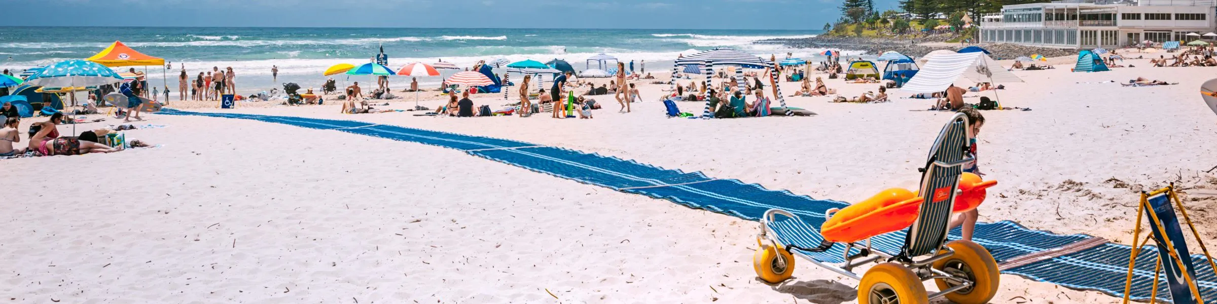 Beach with an access mat leading to the shoreline, a beach wheelchair, and people using umbrellas and tents along the sand.