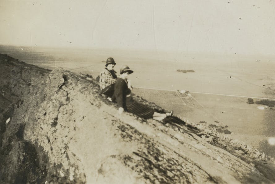 Historic sepia tinted photo of two women sitting on the outer sloped rim of an extinct volcano, with wide flat countryside in the distancewith 