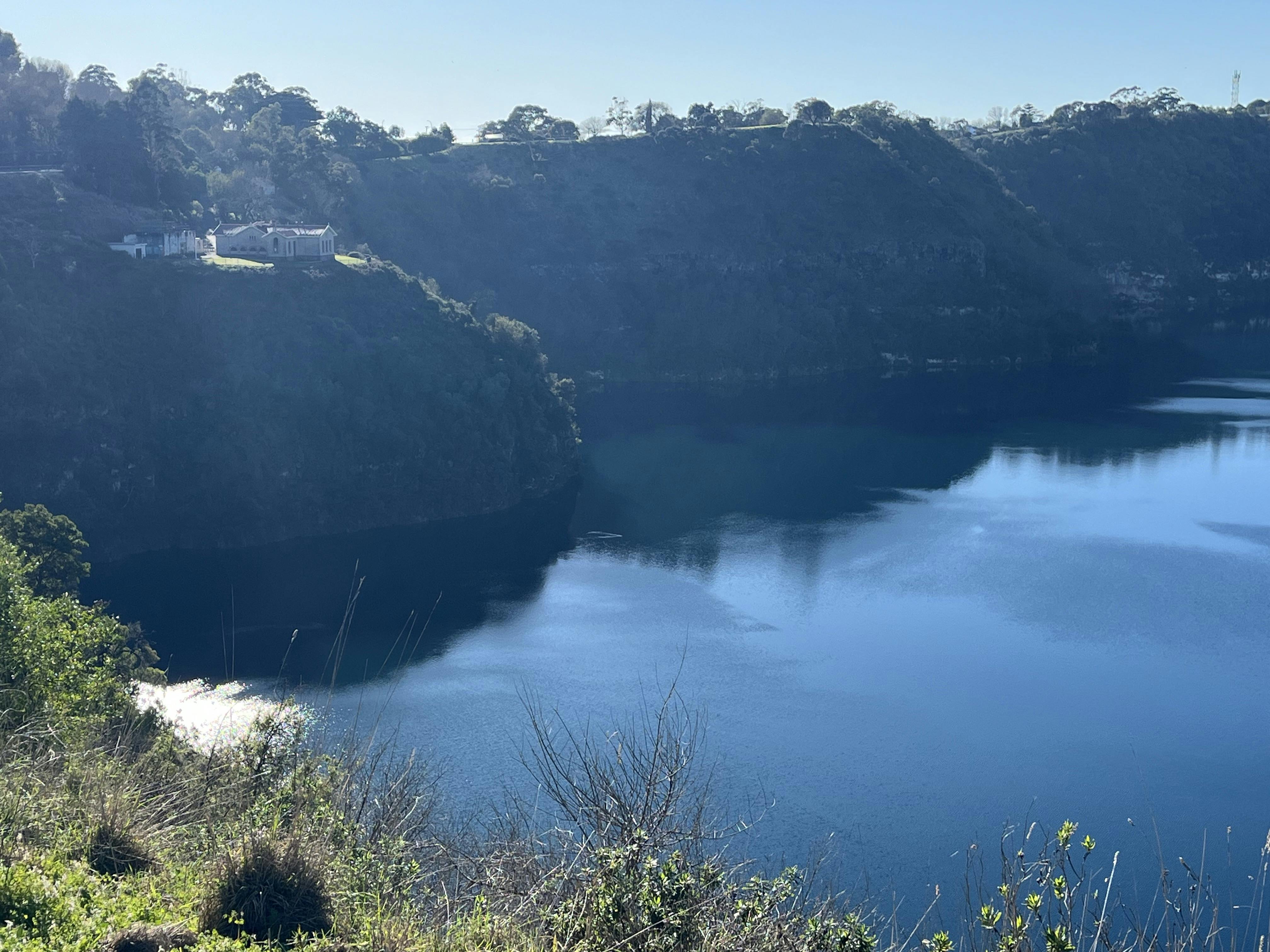 Partial view of crater lake, with blue water and some crater rim including a small cluster of buildings near the edge