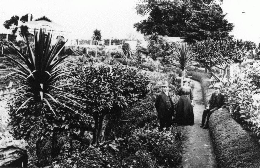 Historic black and white photo of three people in a garden with mature, semi-formal plantings