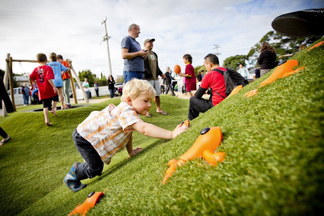 Child playing with toy frogs on the grass while more people practice rugby in the background