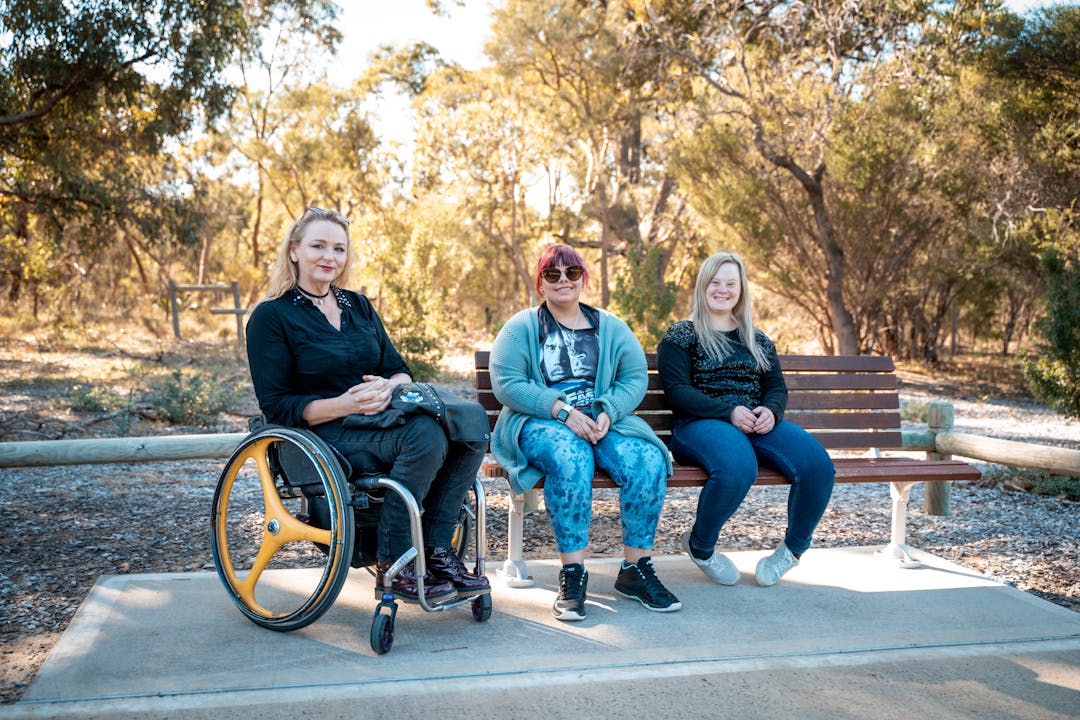 Three community members with disability sitting side by side, one in a wheelchair and two on a park bench.