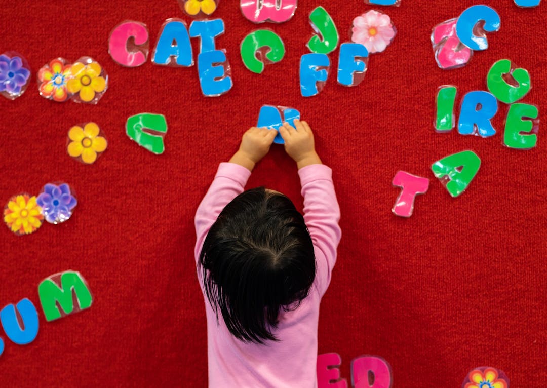 Child sticking coloured letters on a red board with back to camera.