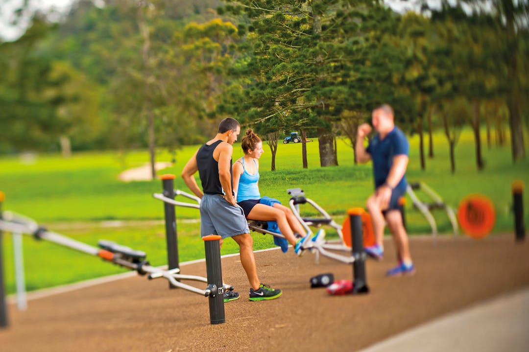 Two fit looking men and one fit looking woman using outdoor exercise equipment in a local park.